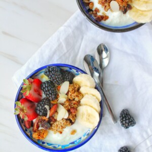 Two blue bowls of yogurt topped with Easy Nutty Cardamom Granola, banana slices, strawberries, blackberries, almonds, and a few nuts sit on a white cloth with two spoons and extra blackberries beside them on a marble surface.