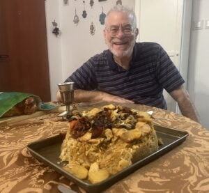 An older man with glasses and a striped shirt smiles at a table set with a large tray of maqluba, a silver goblet, and challah bread. The room features wall decorations and a patterned tablecloth.
