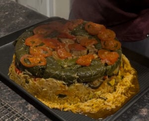 A round dish of maqluba, or makloubeh, an upside-down Middle Eastern rice specialty, topped with cooked grape leaves and sliced tomatoes, with layers of rice and vegetables visible on a baking tray.
