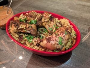A red oval dish filled with spiced baked chicken mandi pieces on a bed of seasoned rice, garnished with chopped herbs and slivered almonds, sits on a wooden table next to a bowl of orange sauce.