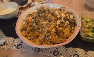 A large bowl of biryani rice with pieces of meat, reminiscent of lamb kabsa, garnished with slivered almonds and herbs, surrounded by side dishes on a wooden table with a patterned table runner.