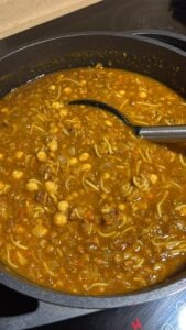A pot of hearty Harira Soup containing chickpeas, noodles, and chunks of meat in a rich, orange-brown broth, with a ladle resting inside the pot.
