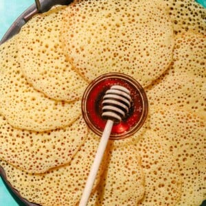 Baghrir placed on a large grey tray with a small bowl of honey in it with a honey dipper on an aqua background with a glass of water in the top corner
