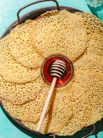 Baghrir placed on a large grey tray with a small bowl of honey in it with a honey dipper on an aqua background with a glass of water in the top corner
