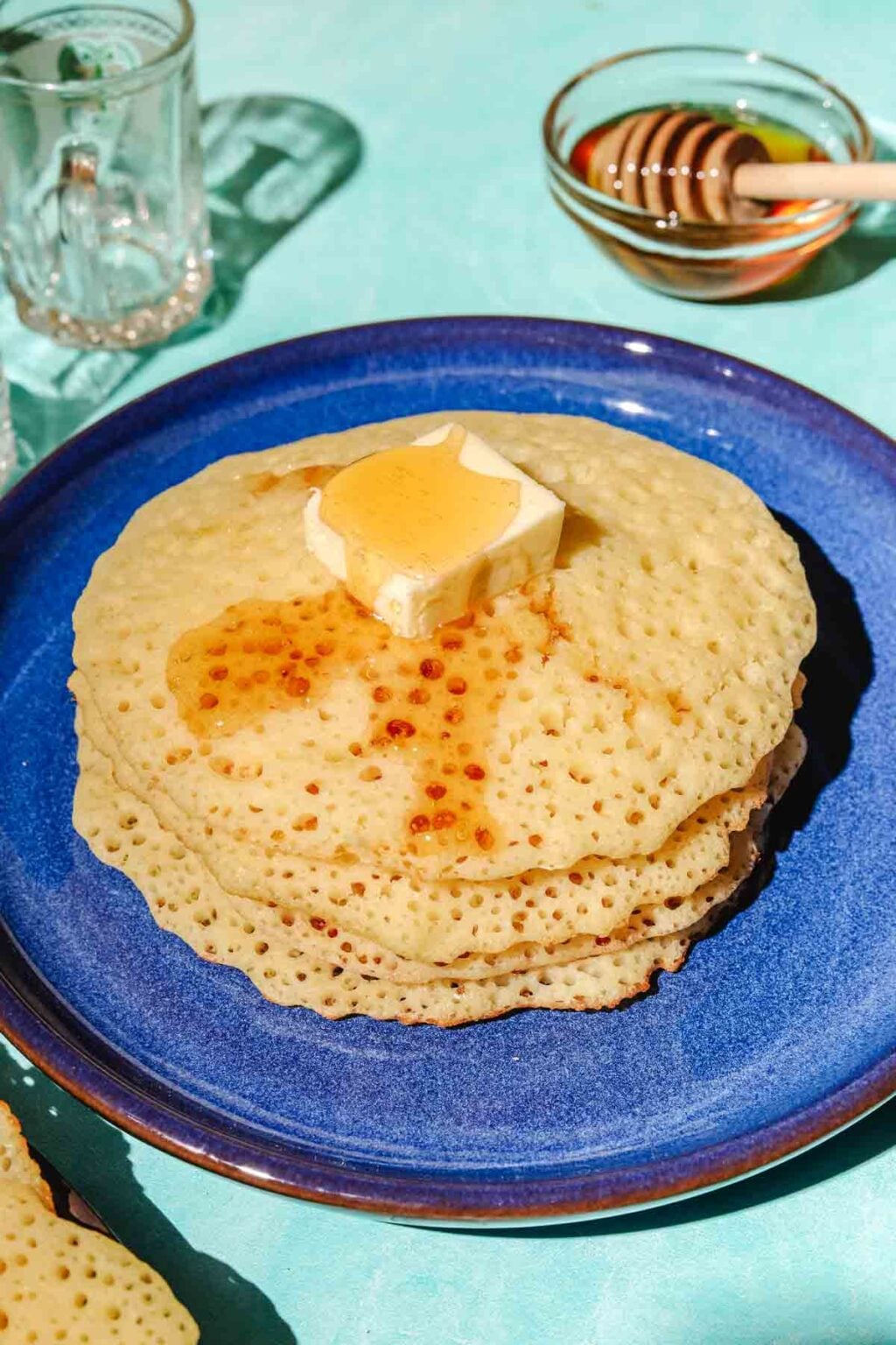 Stacked Baghrir on a dark blue plate with glasses behind it and honey, a bowl of honey with a honey dipper behind it, on an aqua background