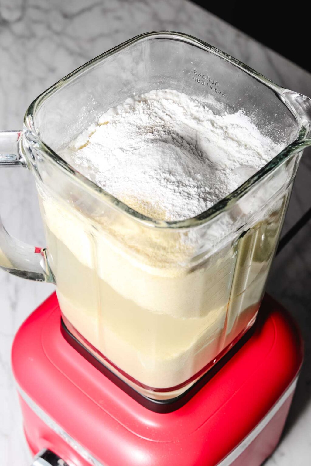 Baghrir batter ingredients, fine semolina, water, yeast, baking powder, sugar and salt, in glass blender jug on a red base on a light marble background