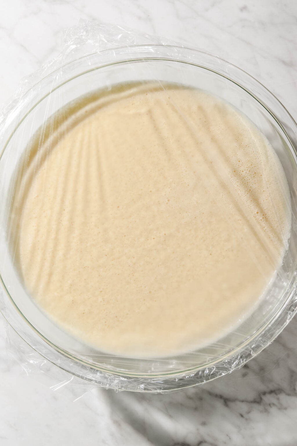Baghrir batter in large glass bowl covered in plastic wrap on light marble background
