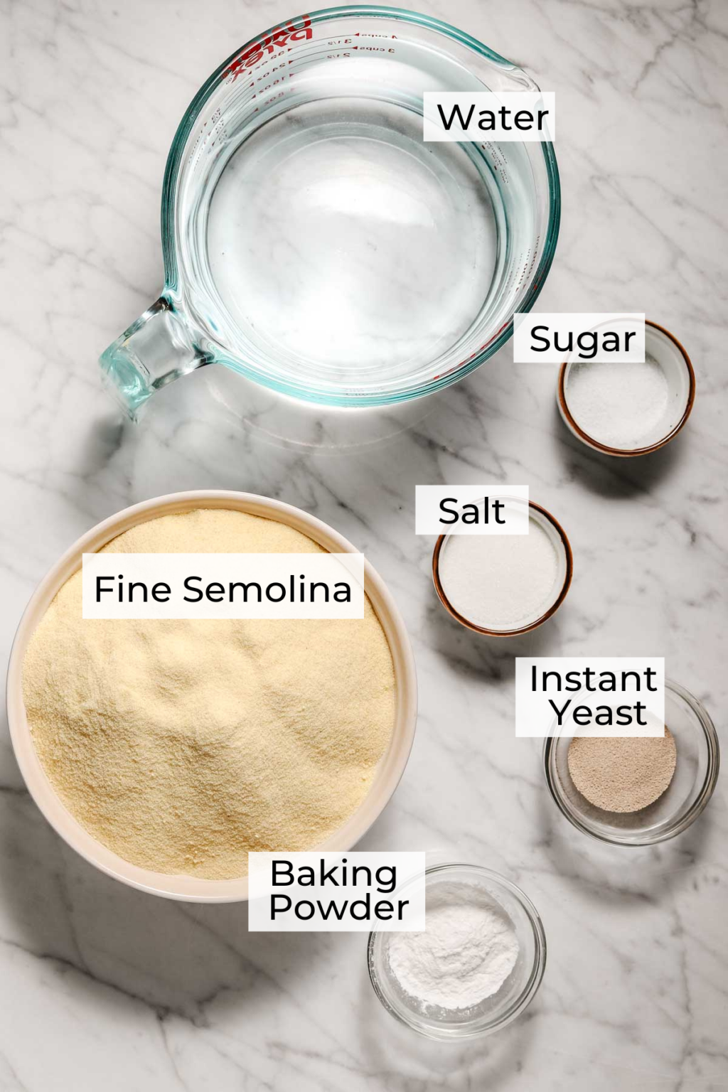 Baghrir ingredient shot with a measuring cup of water, small bowls of sugar, salt, instant yeast and baking powder, medium sized bowl of fine semolina on a light marble background