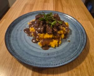 A blue plate with mashed sweet potatoes topped with tender chuck roast, brown gravy, and fresh parsley, placed on a wooden table.