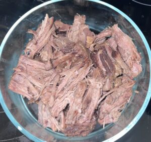 A clear glass bowl filled with shredded, cooked beef from a tender chuck roast sits on a black surface. The meat appears browned, with visible fibers and some pieces darker than others.