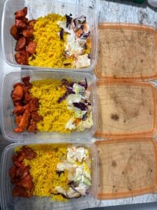 Three plastic meal prep containers filled with yellow rice, pieces of seasoned halal cart chicken, and a portion of mixed salad with cabbage and carrots. The containers have their lids placed beside them.