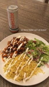 A plate of halal cart chicken with yellow rice, tangy sauces, and fresh salad sits beside a can of “Palestine Cola” on a wooden table. Text reads, we have halal cart at home!.