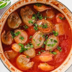 White handled pot with stuffed onions in a tomato broth and chopped parsley on top on a blue and white tiled background with a bowl of chopped parsley at the side.