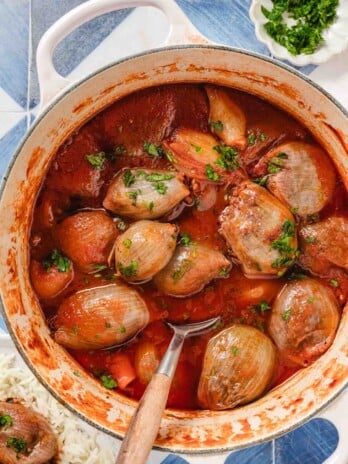 White handled pot with stuffed onions in a tomato broth and chopped parsley on top and a wooden handled serving spoon, a white plate of rice and stuffed onions on top on a blue and white tiled background with a bowl of chopped parsley at the side.