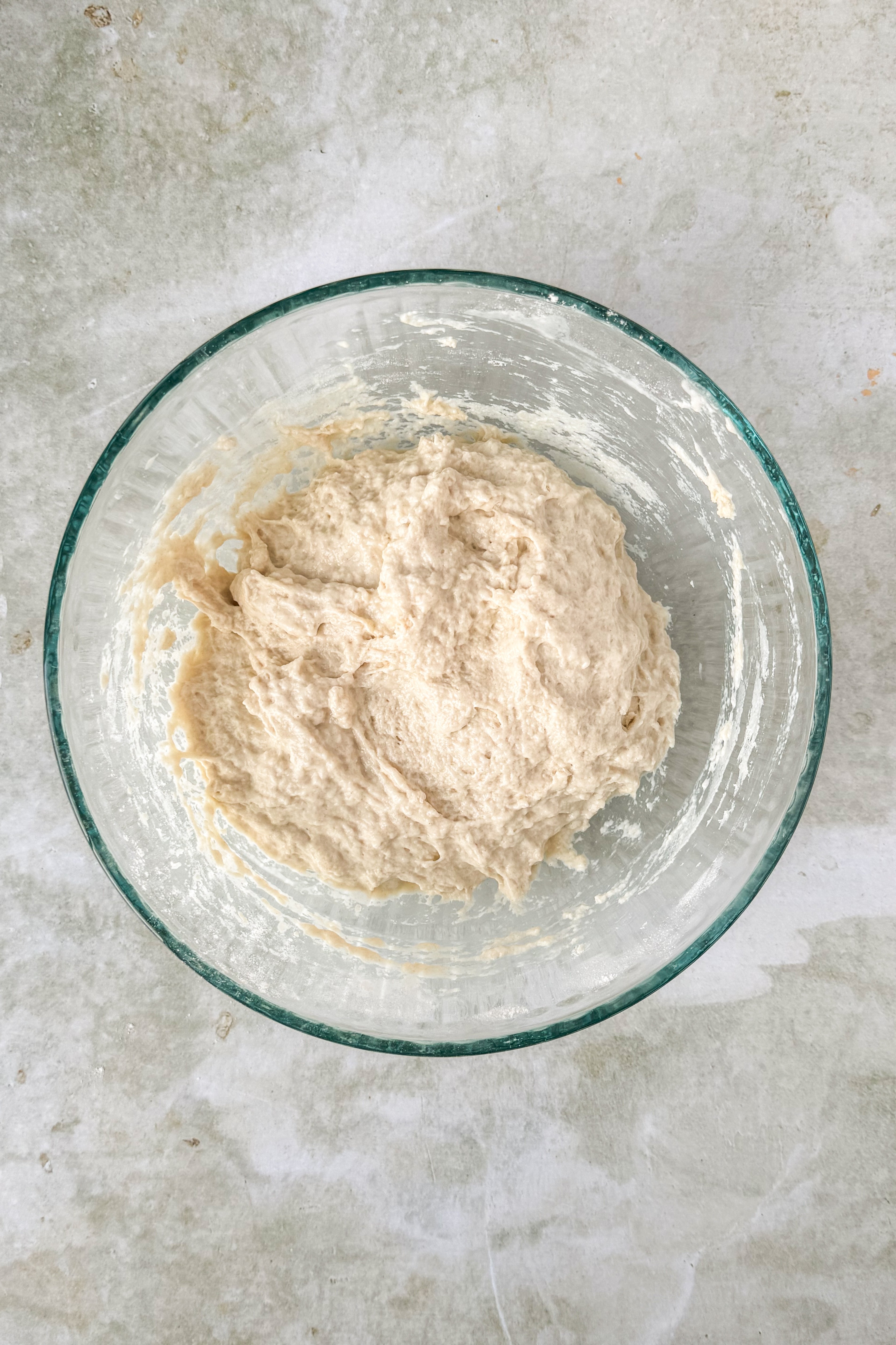 Wet dough in a glass bowl on an olive green marbled background.