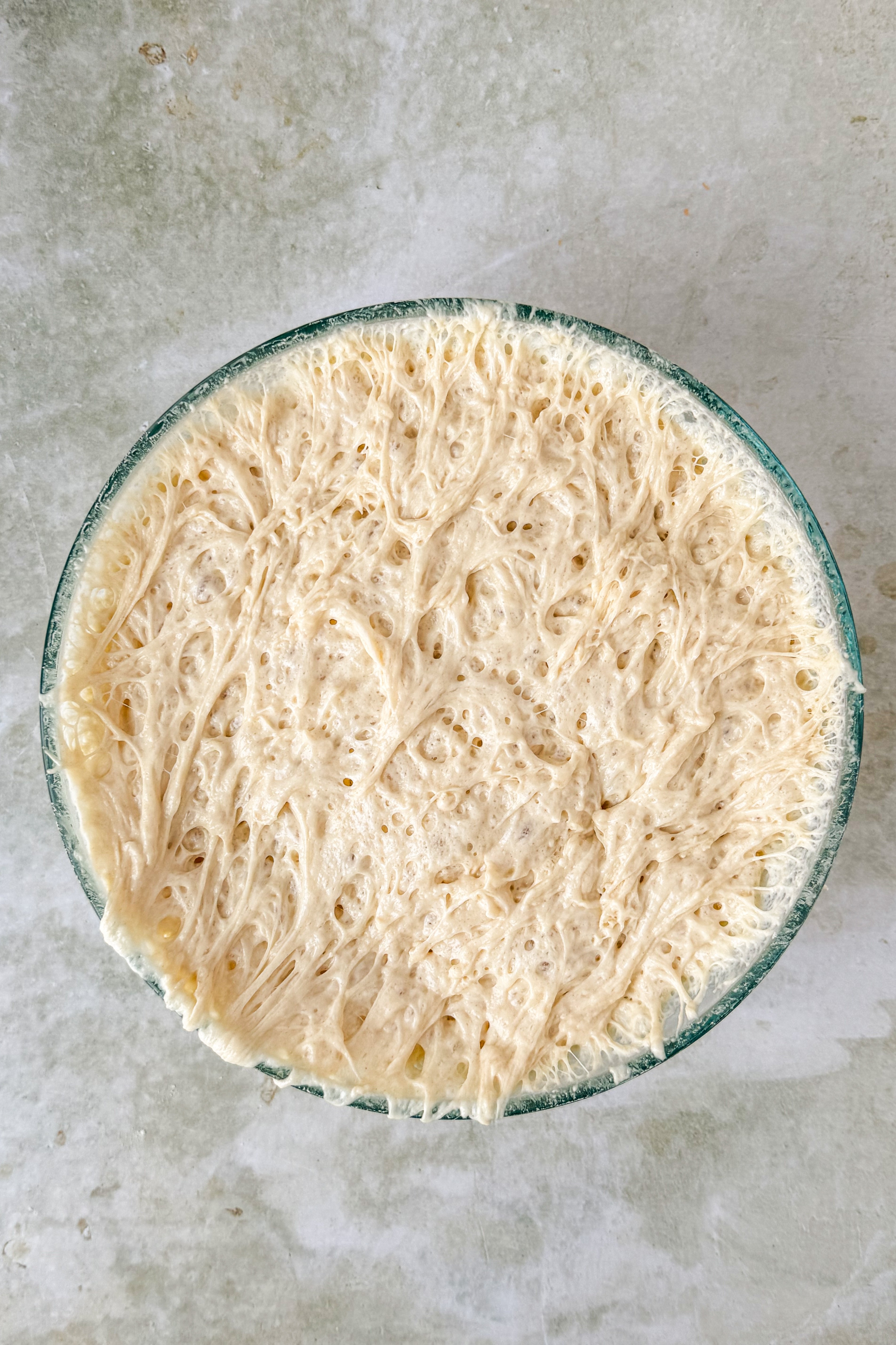 Risen wet dough in a glass bowl on an olive green marble background.