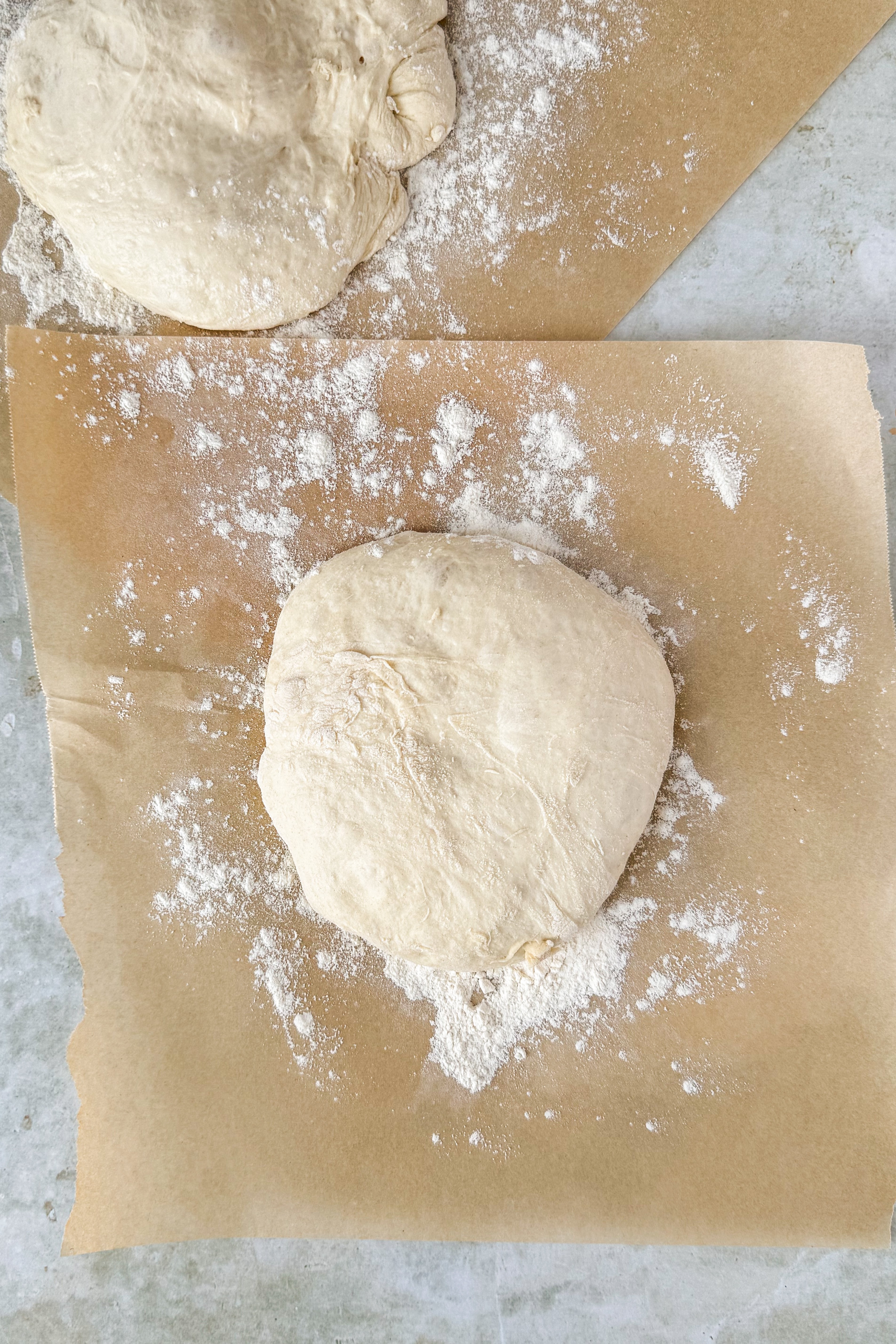 Two dough balls on floured parchment paper on an olive marble background.