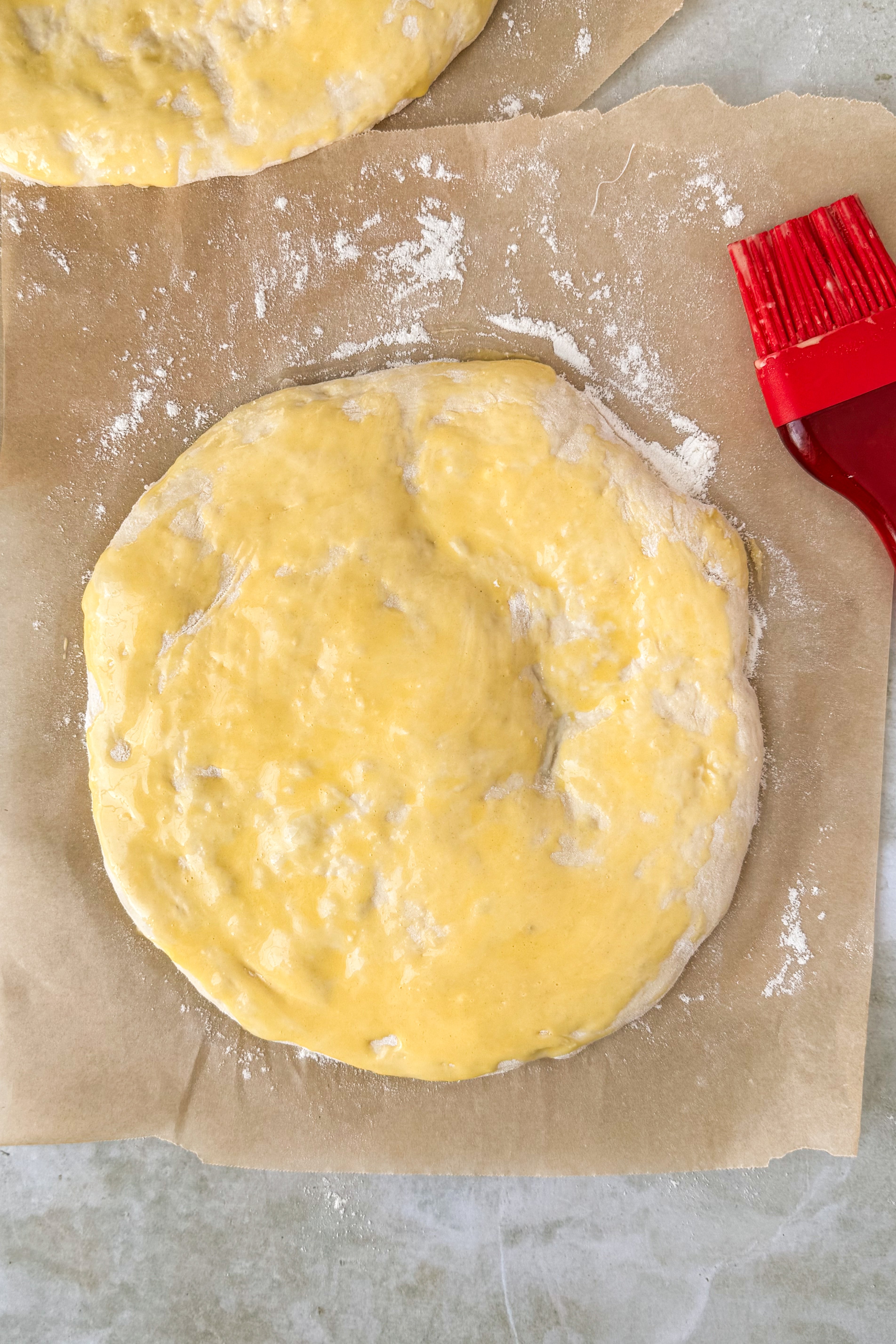 Two dough circles on floured parchment paper brushed with egg wash with a red silicon brush at the side on an olive marble background.