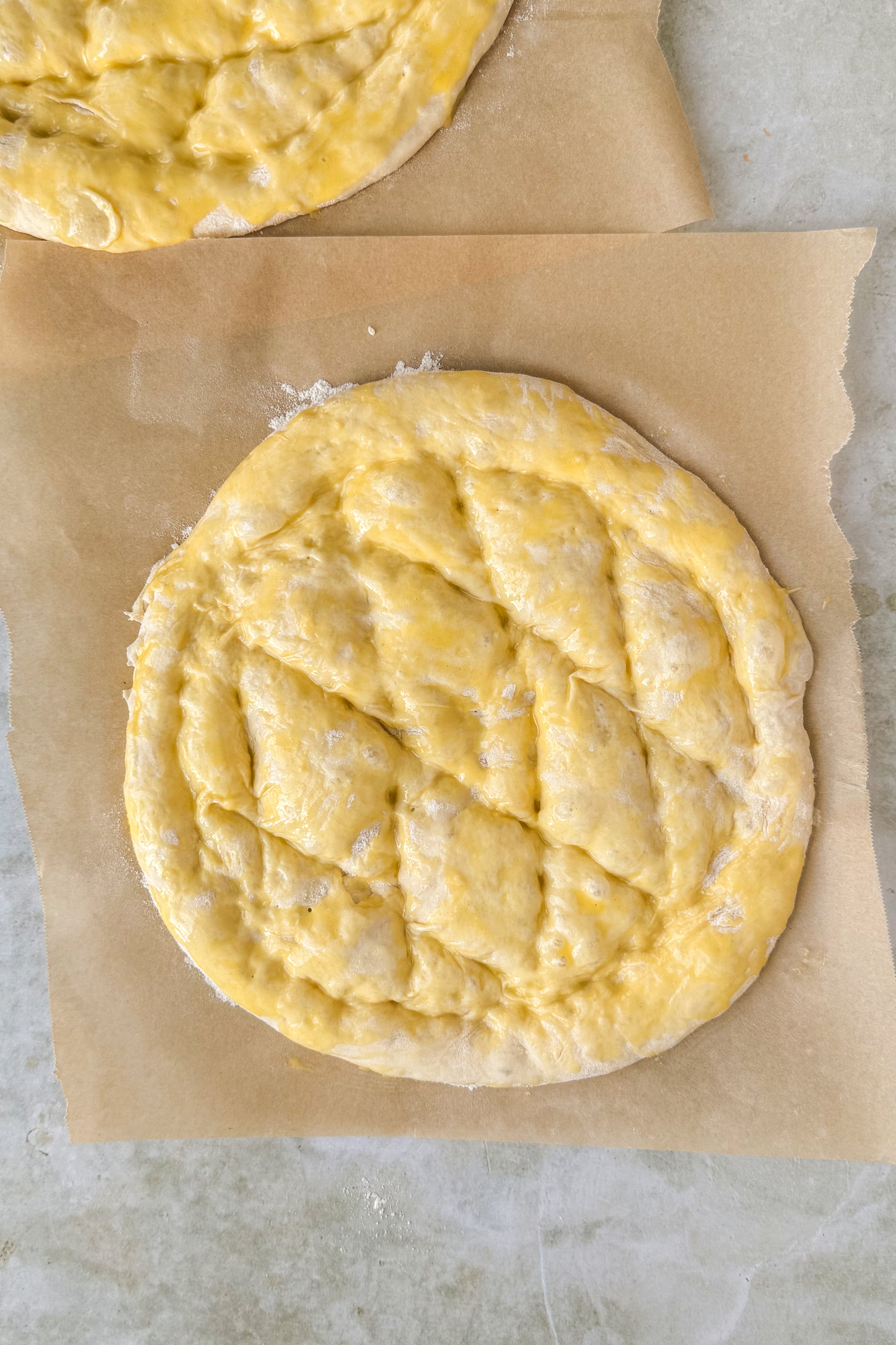 Two dough circles on floured parchment paper brushed with egg wash with lines pressed onto the dough in a diamond shape on an olive marble background.
