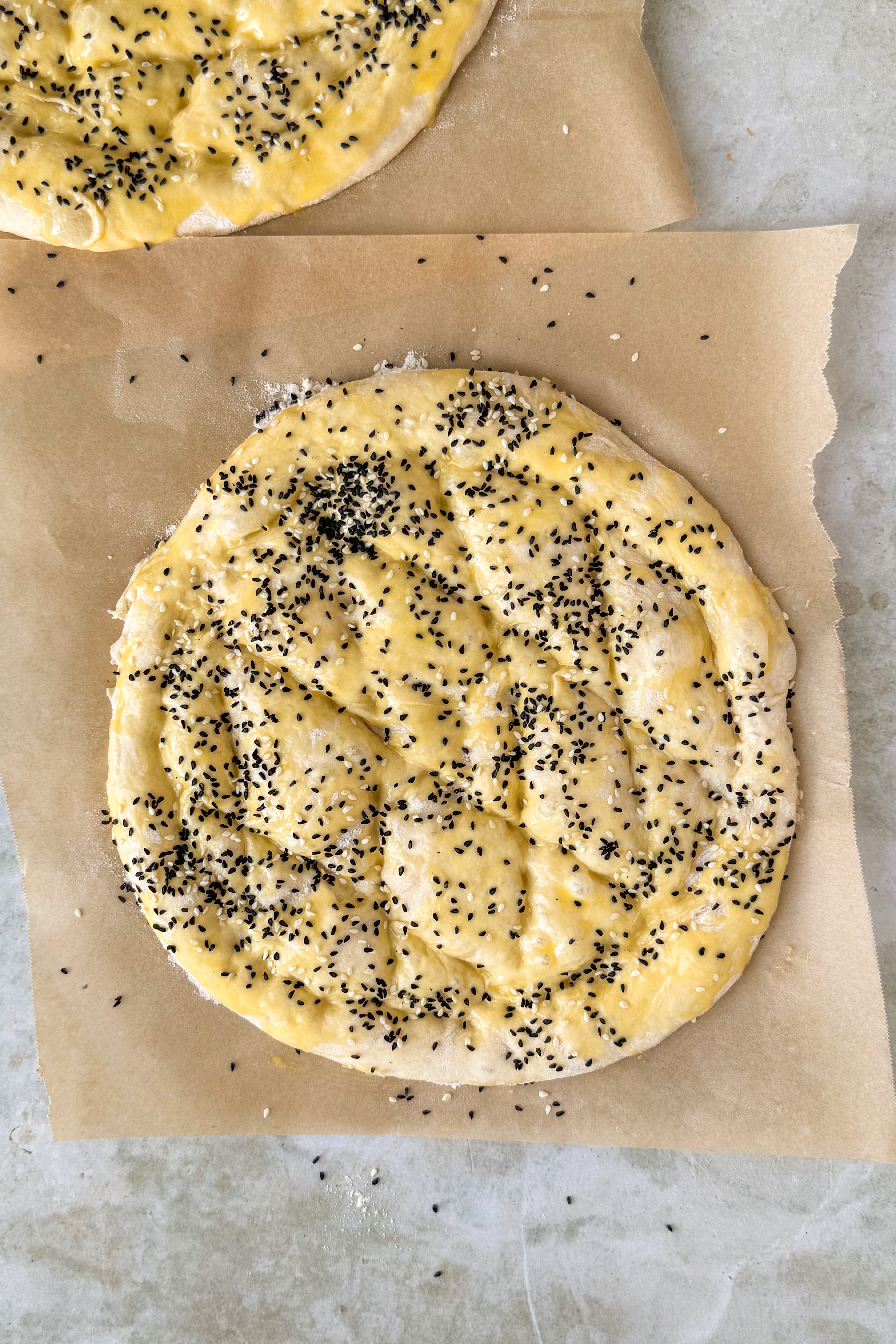 Two dough circles on floured parchment paper brushed with egg wash with lines pressed onto the dough in a diamond shape and nigella seeds and sesame seeds sprinkled on top on an olive marble background.