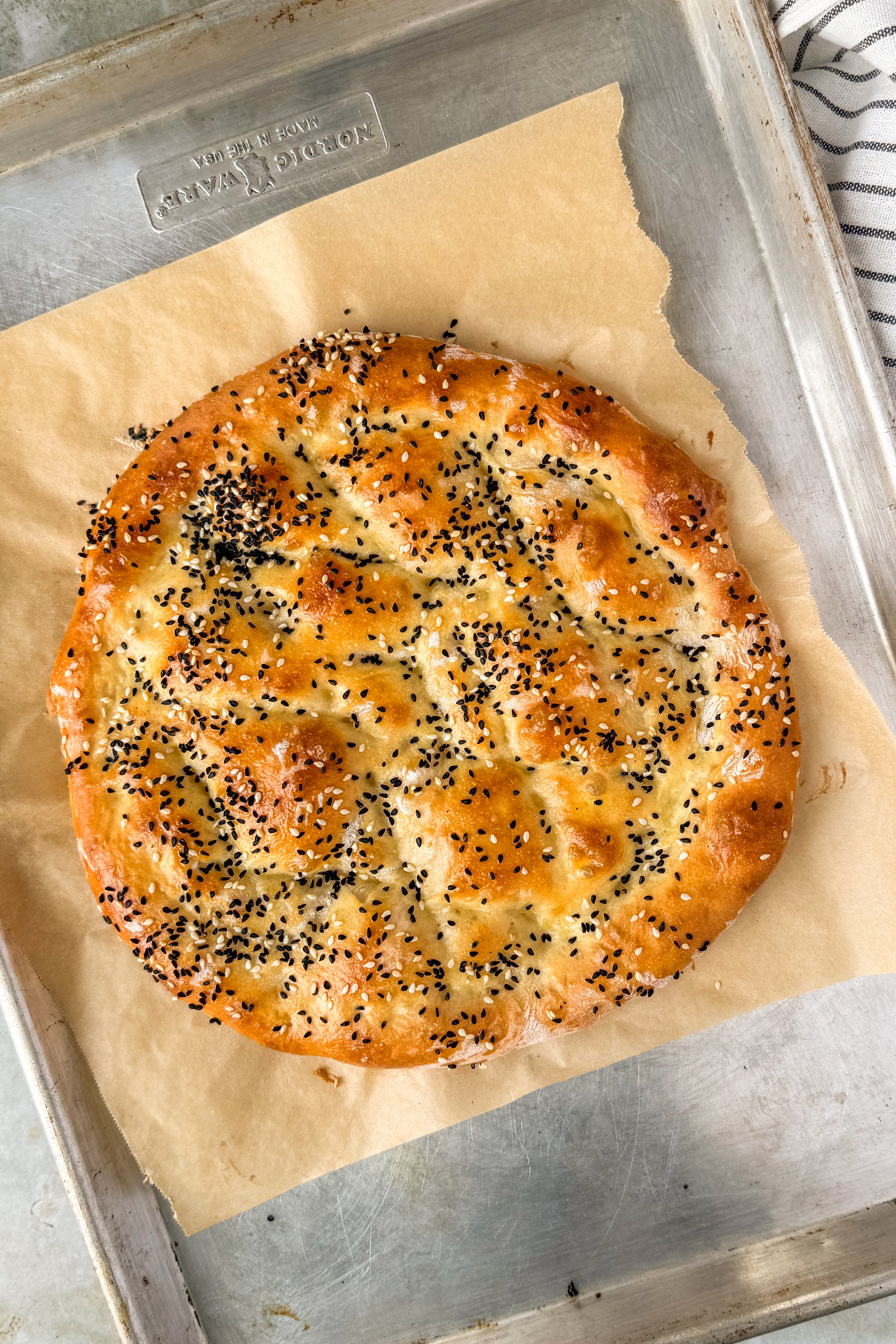 Baked round flatbread with nigella seeds and sesame seeds on a piece of parchment on a baking tray with a black and white towel at the side.