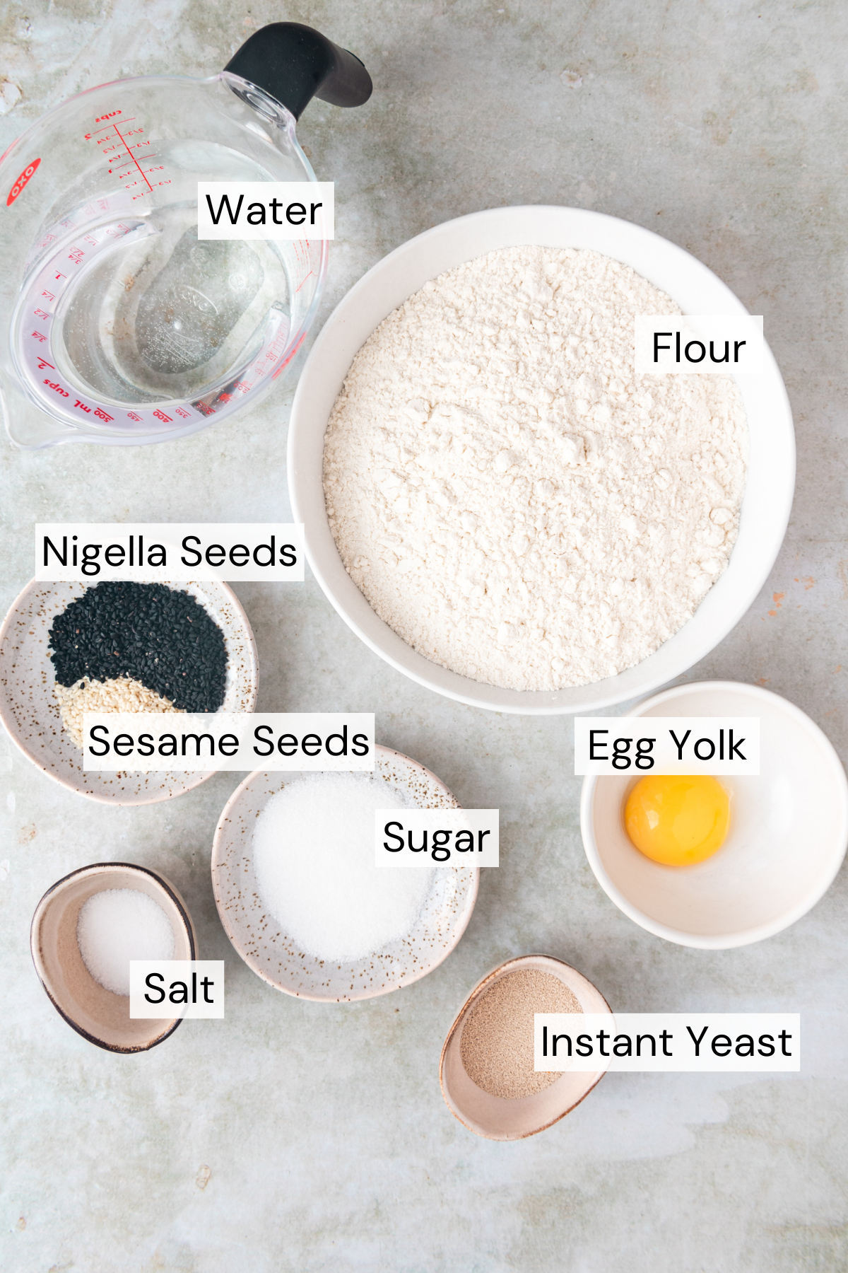 Ingredient shot with a measuring cup with water, a white bowl with flour, a small plate with nigella seeds and sesame seeds, a small plate with sugar, a small bowl of salt, a white bowl with egg yolk, and a small bowl of yeast on an olive green marble background.