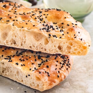 Two sliced flatbreads showing the side view sprinkled with sesame seeds and nigella seeds on a beige marble background with a green glass filled with water behind it, parsley, black olives and tomatoes.