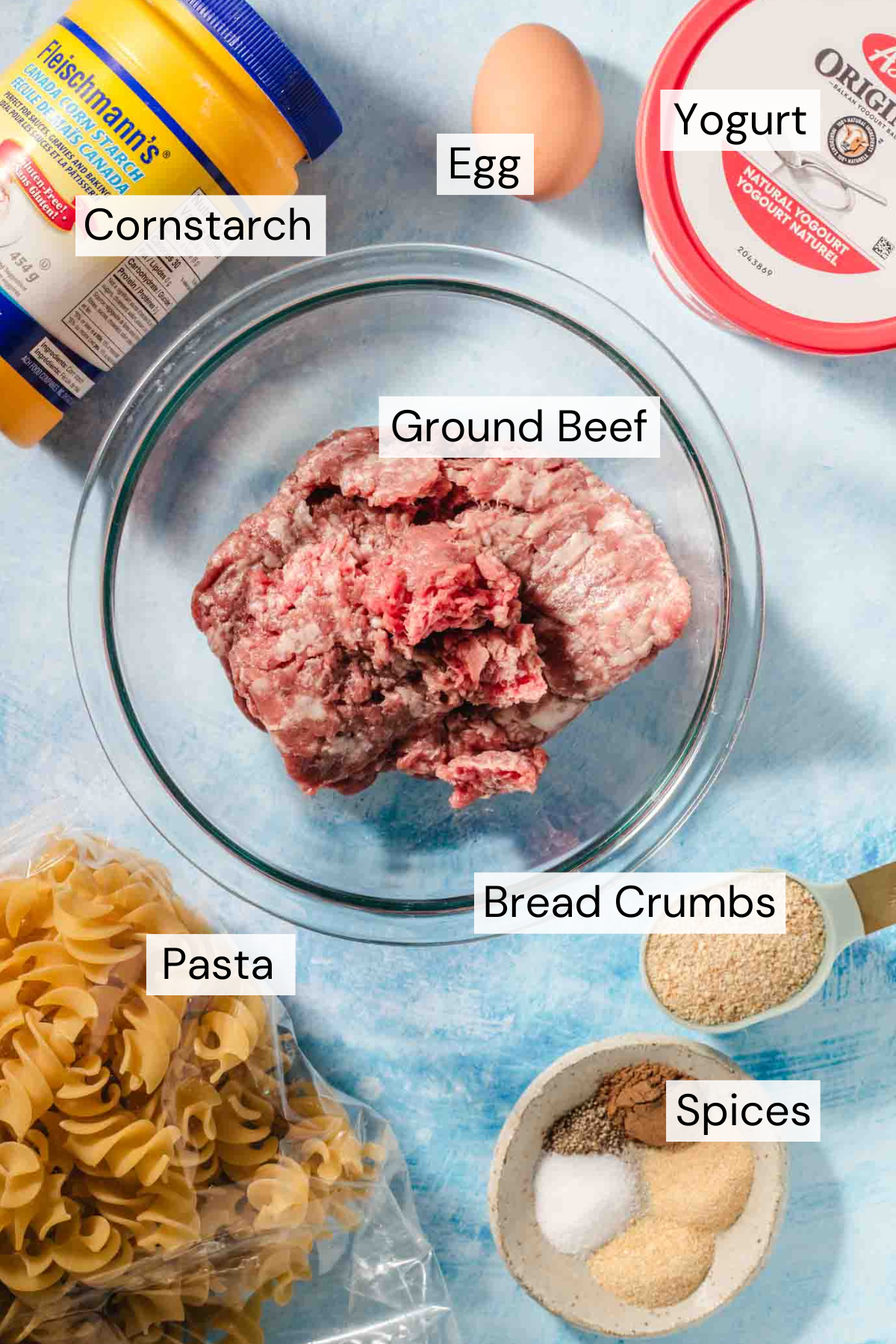 Ingredient shot with a container with a red lid of yogurt, a brown egg, a yellow and blue container of cornstarch, a glass bowl with ground beef, a clear plastic package with spiral pasta, a measuring spoon with bread crumbs and a bowl with spices on a blue background.