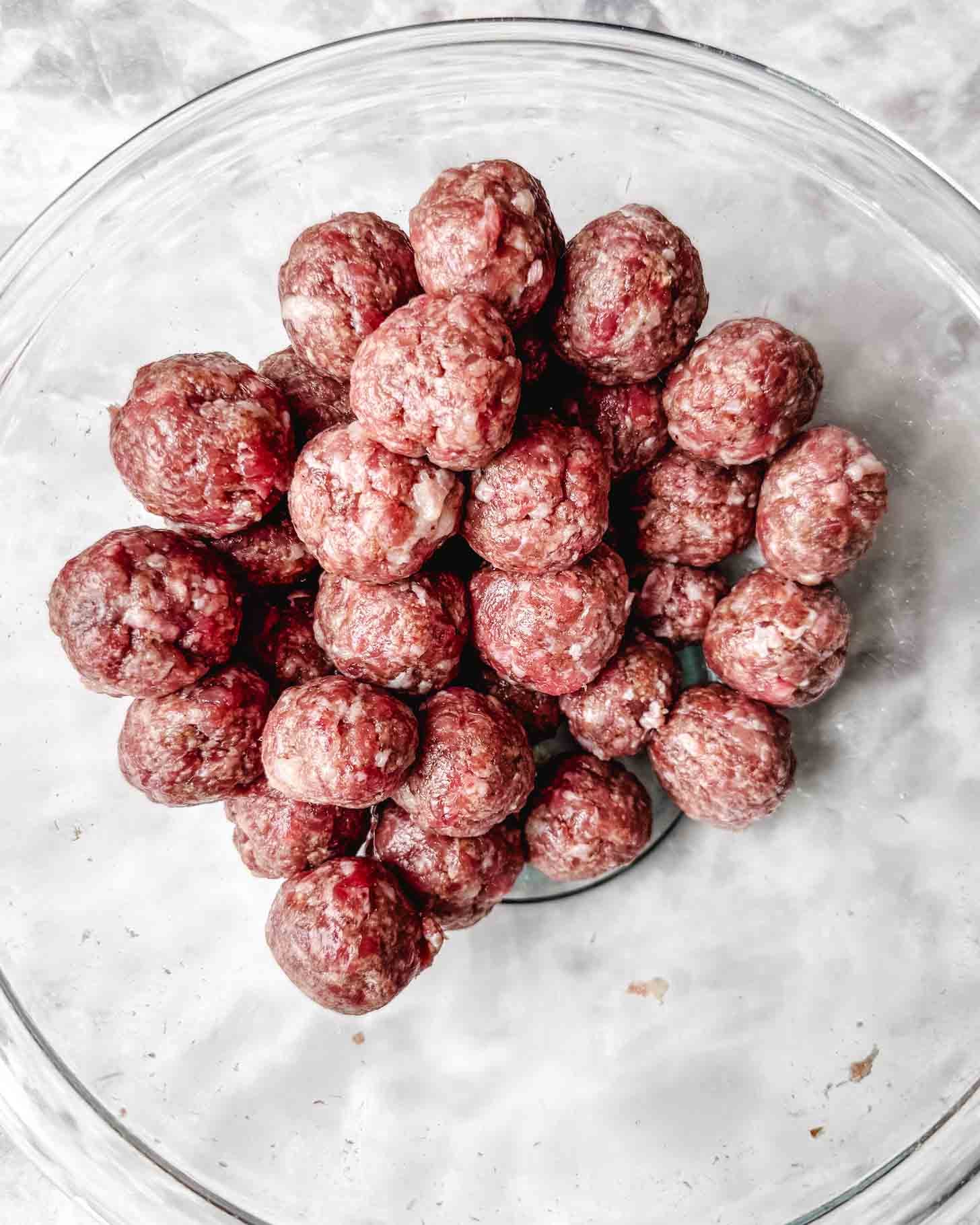 Round brown uncooked meatballs piled into a glass bowl on a marble surface.