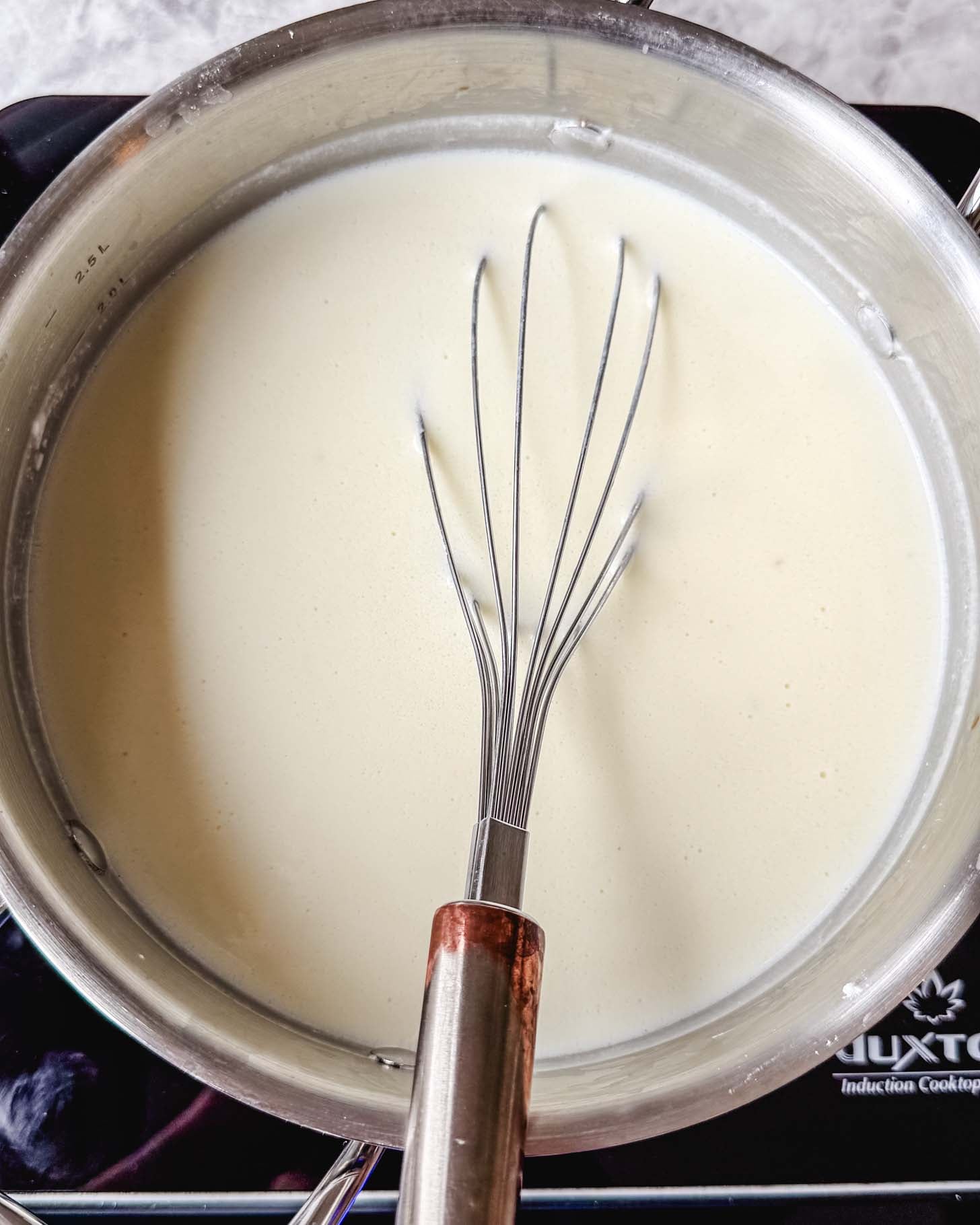 Steel pot with a white creamy liquid with a metal whisk inside on a marble surface.