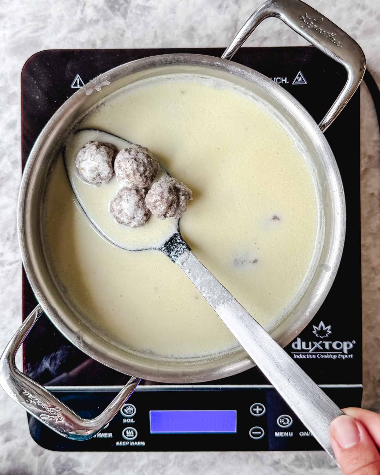 Steel pot with a creamy off white liquid and small brown meatballs in a spoon being held by a hand on a marble background.