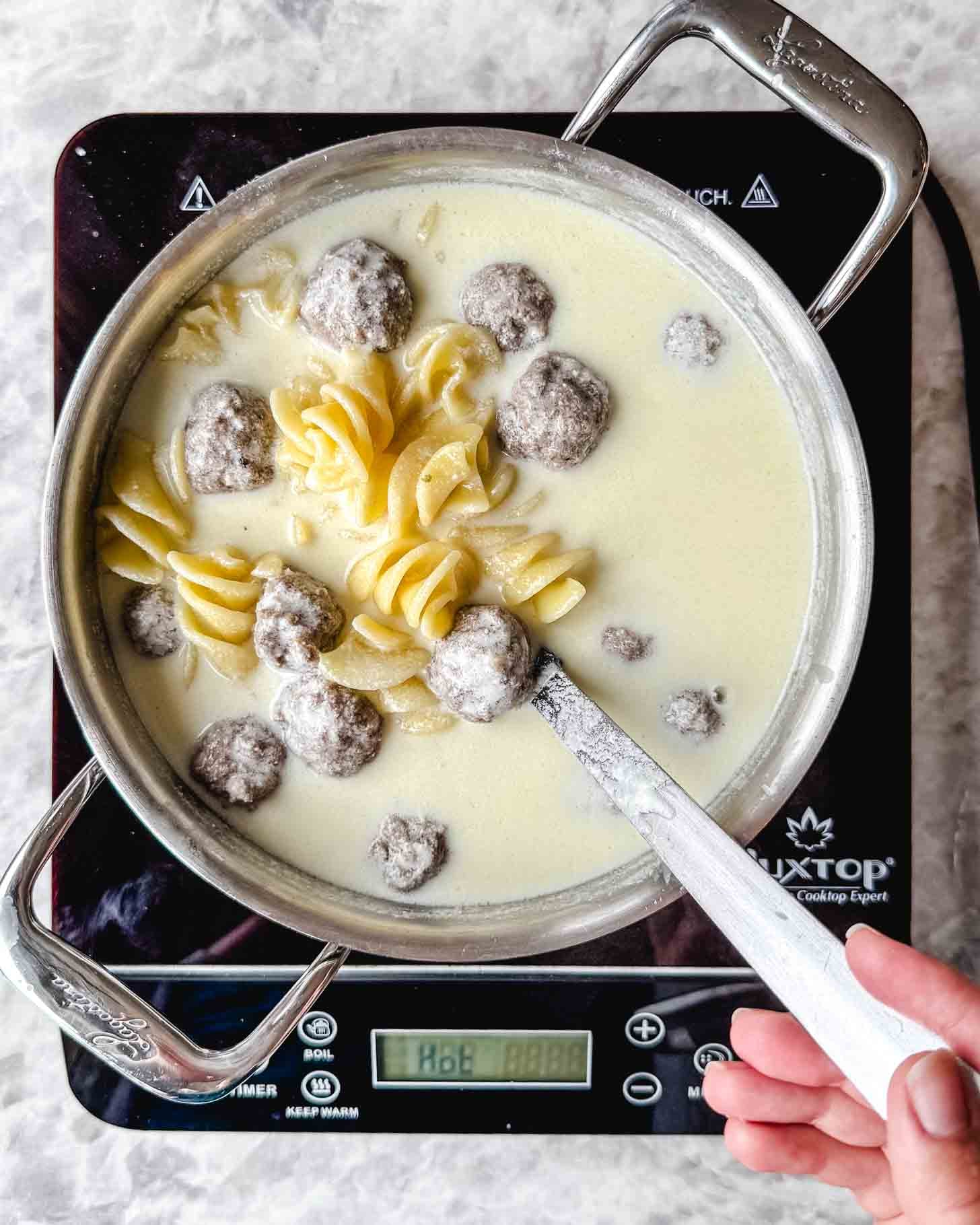 Steel pot with a creamy off white liquid, pasta spirals and small brown meatballs in a spoon being held by a hand on a marble background.