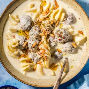 Beige speckled bowl with a creamy off white liquid, small brown meatballs covered in the off white liquid, spiral shaped white pasta and golden toasted pine nuts on top with a metal spoon in the bowl on a blue background with a blue tea towel in the corner and a small blue bowl with toasted pine nuts.