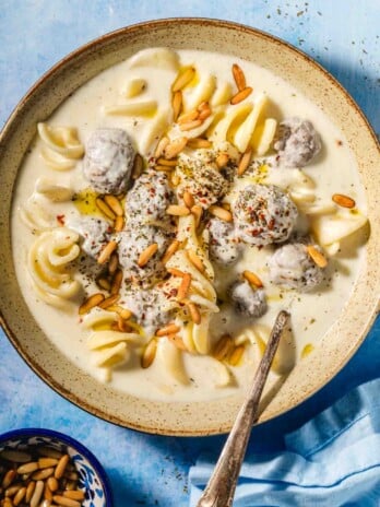 Beige speckled bowl with a creamy off white liquid, small brown meatballs covered in the off white liquid, spiral shaped white pasta and golden toasted pine nuts on top with a metal spoon in the bowl on a blue background with a blue tea towel in the corner and a small blue bowl with toasted pine nuts.