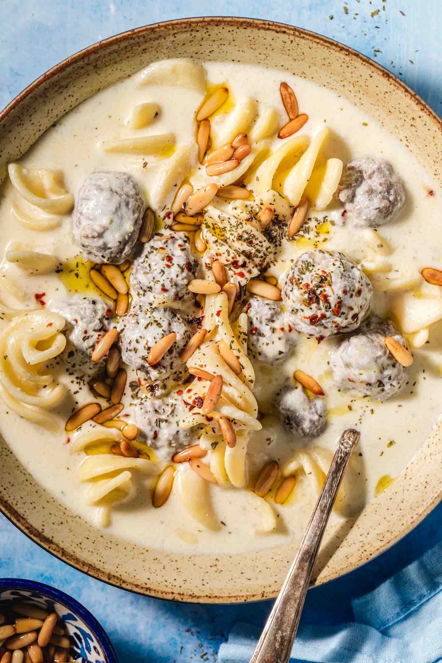 Beige speckled bowl with a creamy off white liquid, small brown meatballs covered in the off white liquid, spiral shaped white pasta and golden toasted pine nuts on top with a metal spoon in the bowl on a blue background with a blue tea towel in the corner and a small blue bowl with toasted pine nuts.