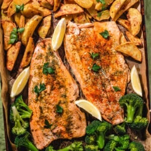 A baking tray with two seasoned salmon fillets, roasted potato wedges, steamed broccoli, and lemon wedges, all garnished with fresh parsley on parchment paper.