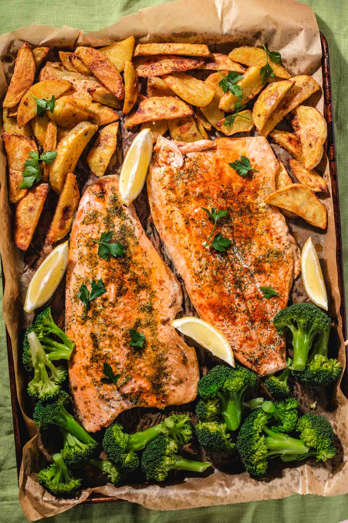 A baking tray with two seasoned salmon fillets, roasted potato wedges, steamed broccoli, and lemon wedges, all garnished with fresh parsley on parchment paper.