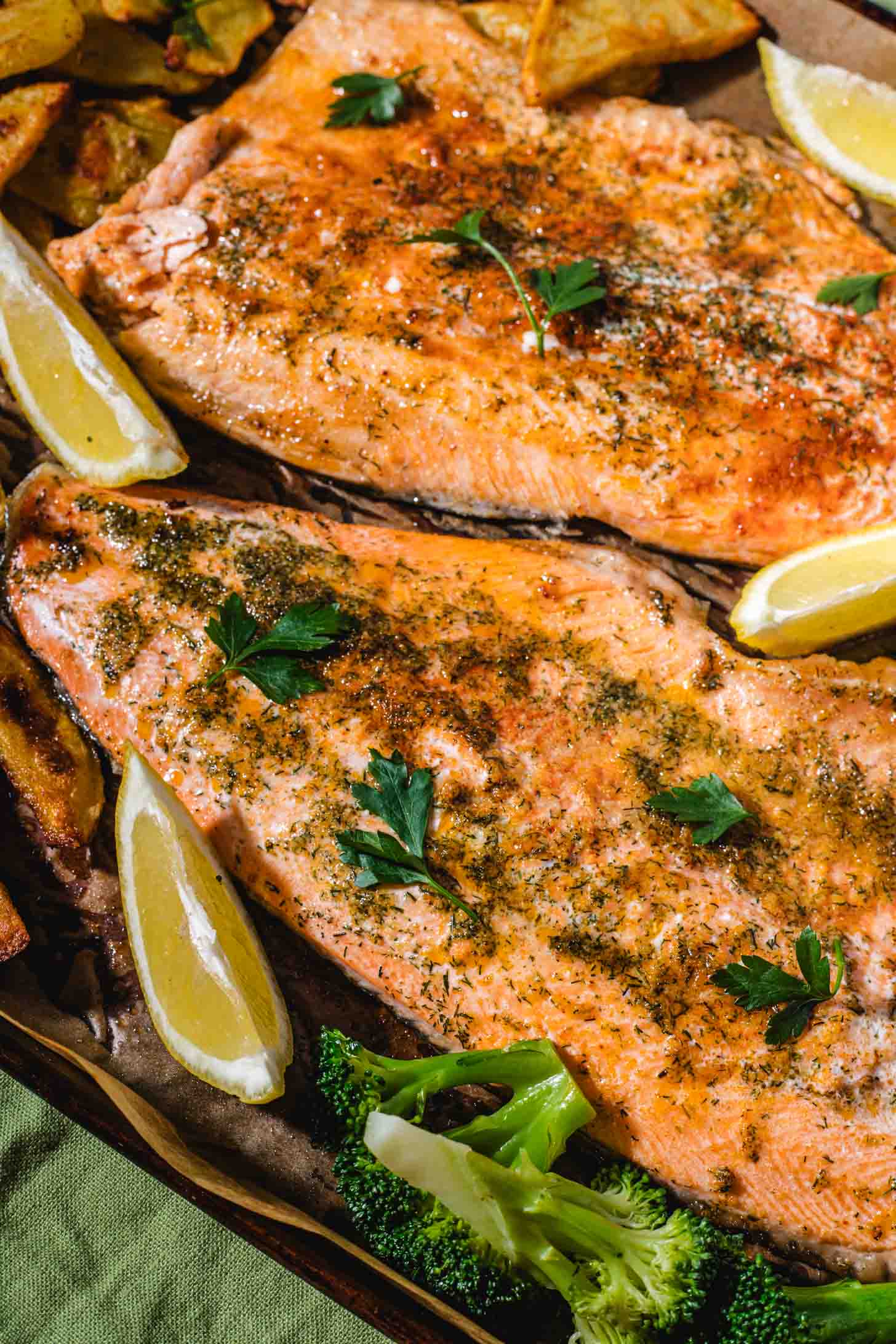 Two baked salmon fillets garnished with herbs and parsley, surrounded by lemon wedges, potato slices, and broccoli on a baking tray.