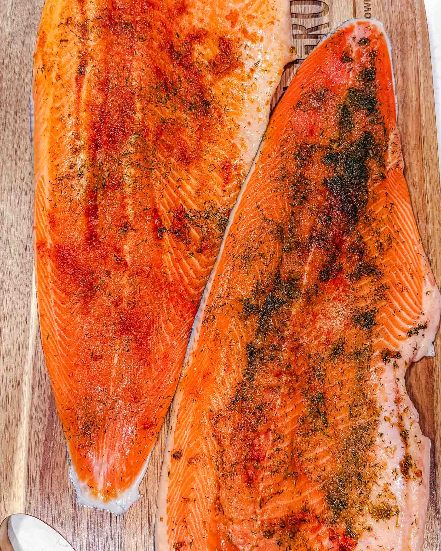 Two raw salmon fillets on a wooden cutting board, seasoned with various spices and herbs, showing bright orange flesh and visible seasoning.