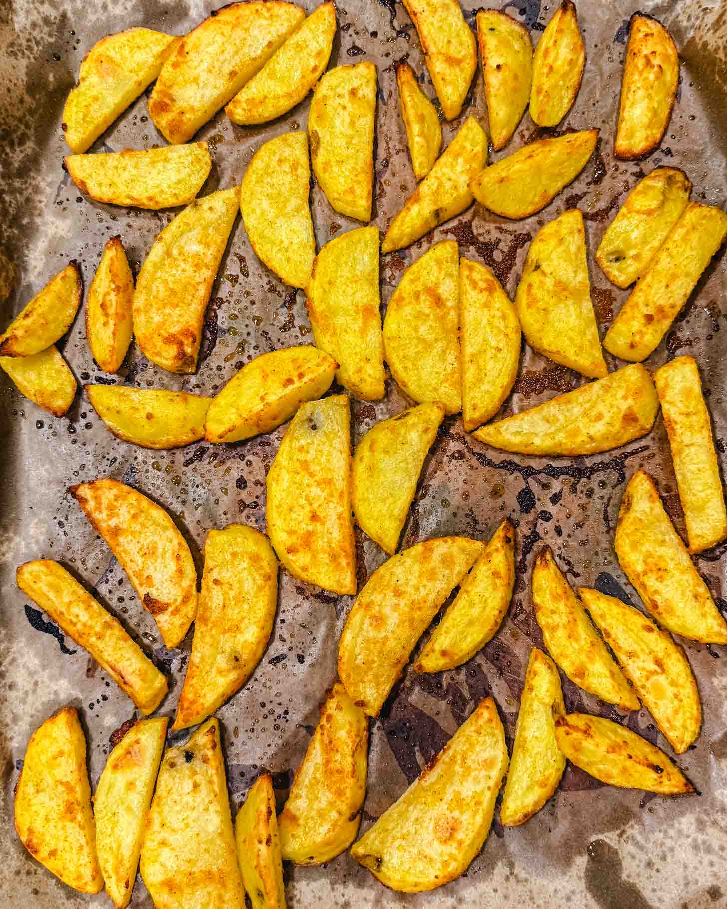 Golden, crispy potato wedges are spread out on a baking sheet lined with parchment paper, showing some browning and oily spots from roasting.