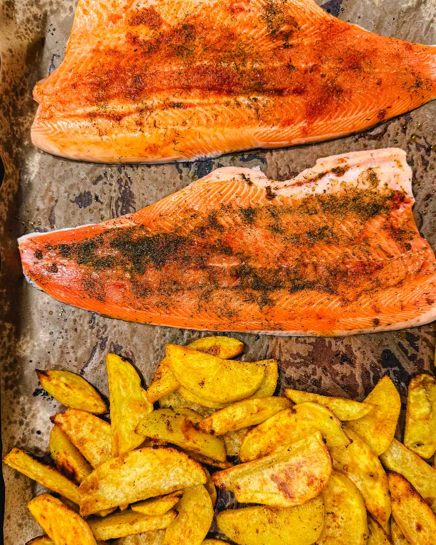 Two fillets of seasoned salmon on a baking sheet with a portion of golden, roasted potato wedges beside them.