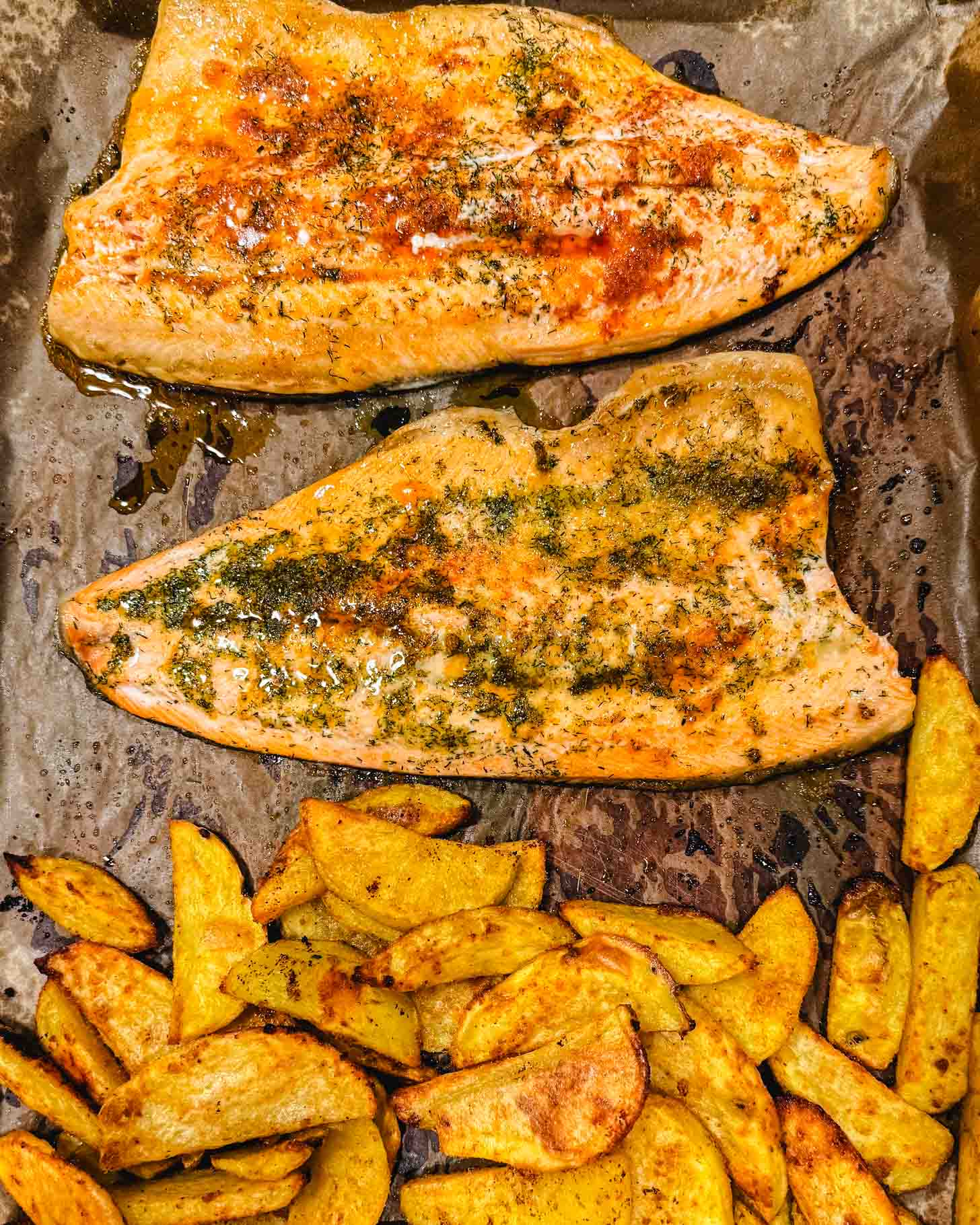 Two seasoned salmon fillets and a pile of golden roasted potato wedges on a parchment-lined baking tray, fresh from the oven.