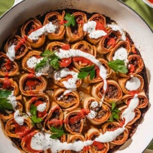 A round pan filled with rolled pastries stuffed with a meat mixture, garnished with fresh parsley, and drizzled with white and red sauces, set on a green tablecloth.