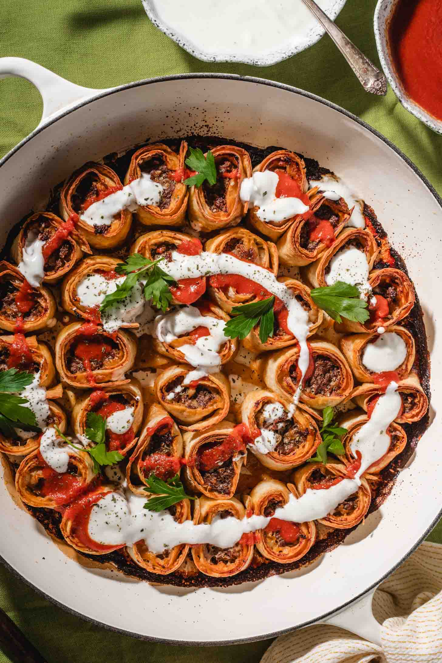 A round pan filled with rolled pastries stuffed with a meat mixture, garnished with fresh parsley, and drizzled with white and red sauces, set on a green tablecloth.