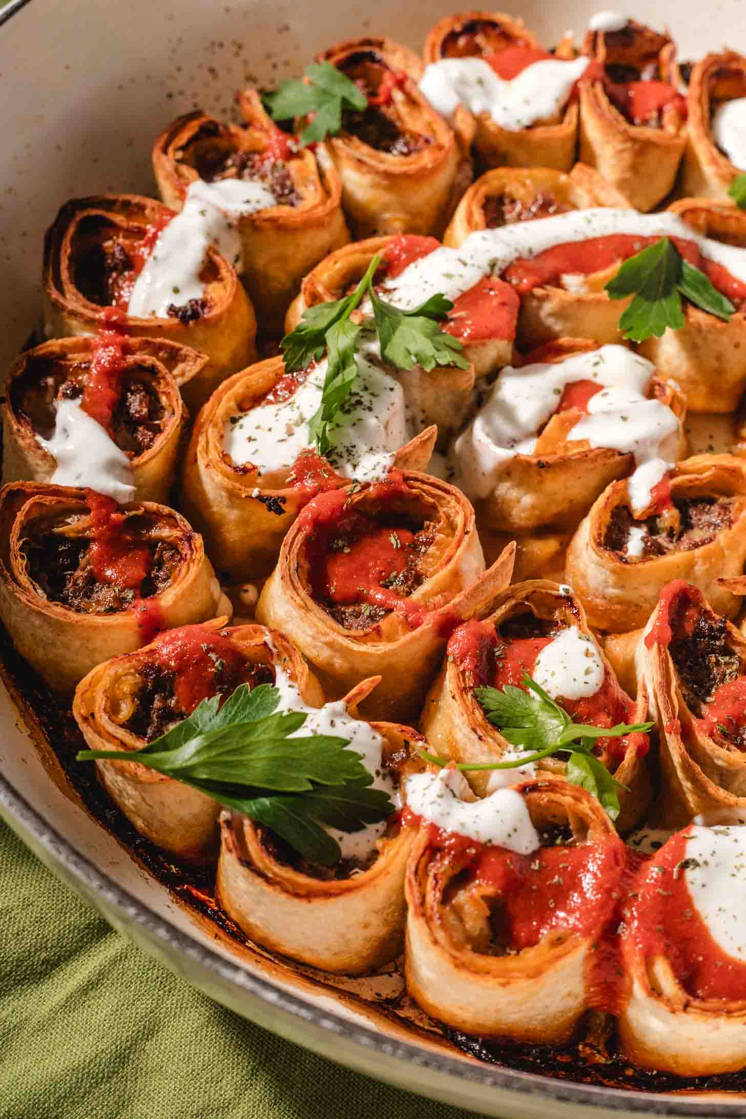 A close-up of rolled tortillas filled with a savory mixture, baked in a pan, and topped with dollops of sour cream, red sauce, and garnished with fresh parsley.