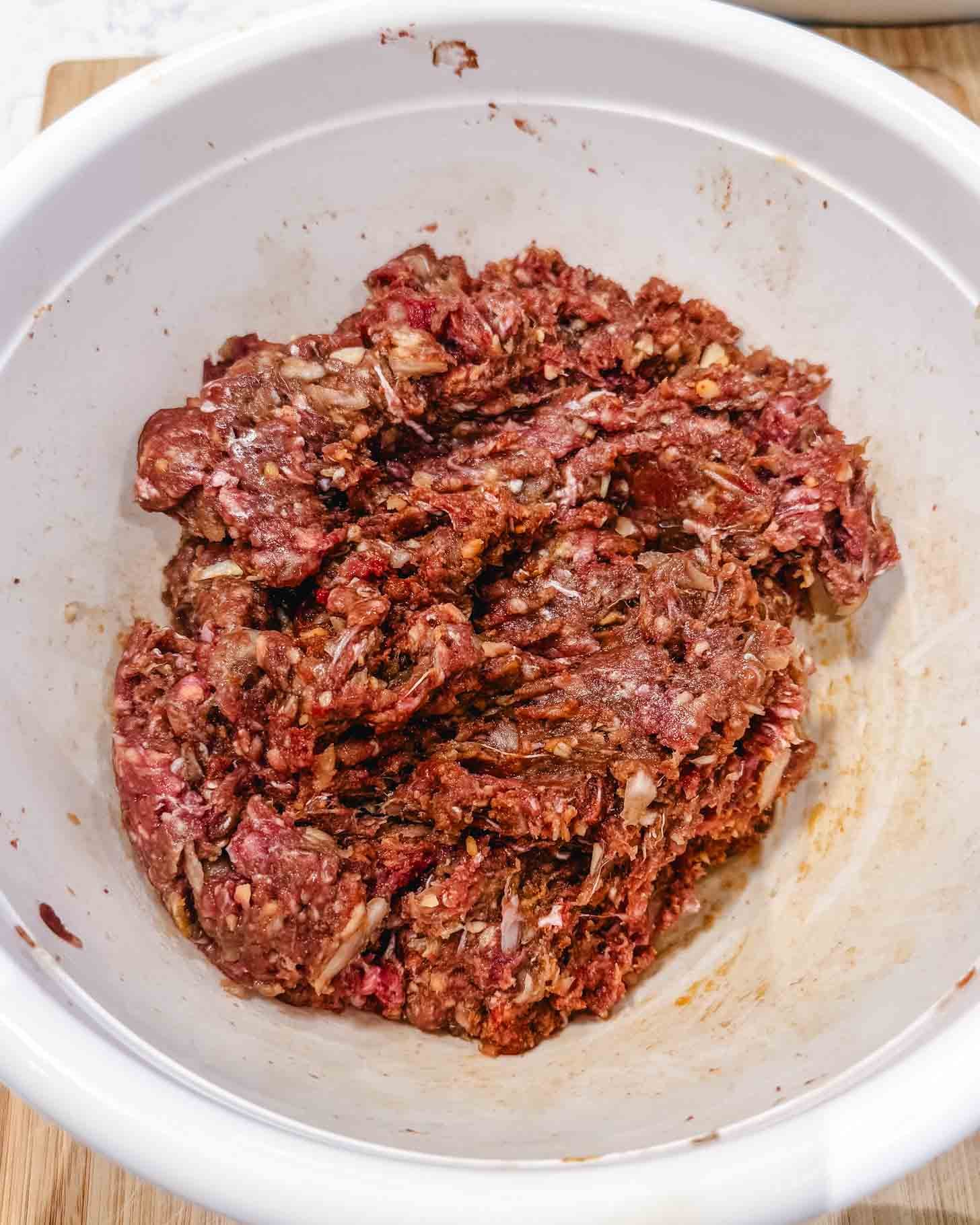 Raw ground beef mixed with seasonings and oats in a white mixing bowl, ready to be used for a recipe such as meatloaf or meatballs.