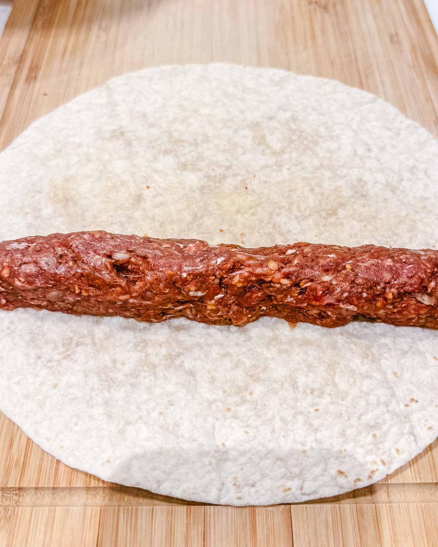 A raw ground meat mixture shaped into a log is placed on a round flour tortilla on a wooden surface, ready to be rolled up.
