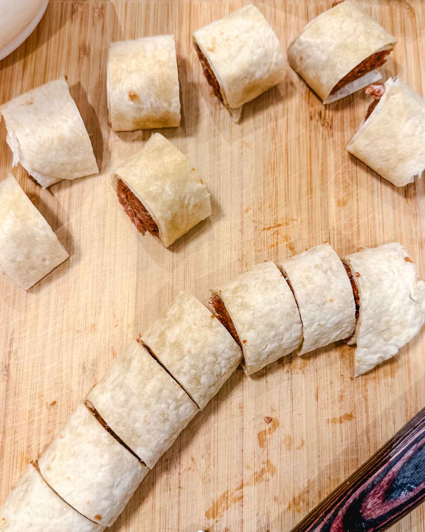 A sliced tortilla wrap with a brown filling is laid on a wooden cutting board. Several bite-sized pieces are separated, showing the rolled wrap and filling inside.