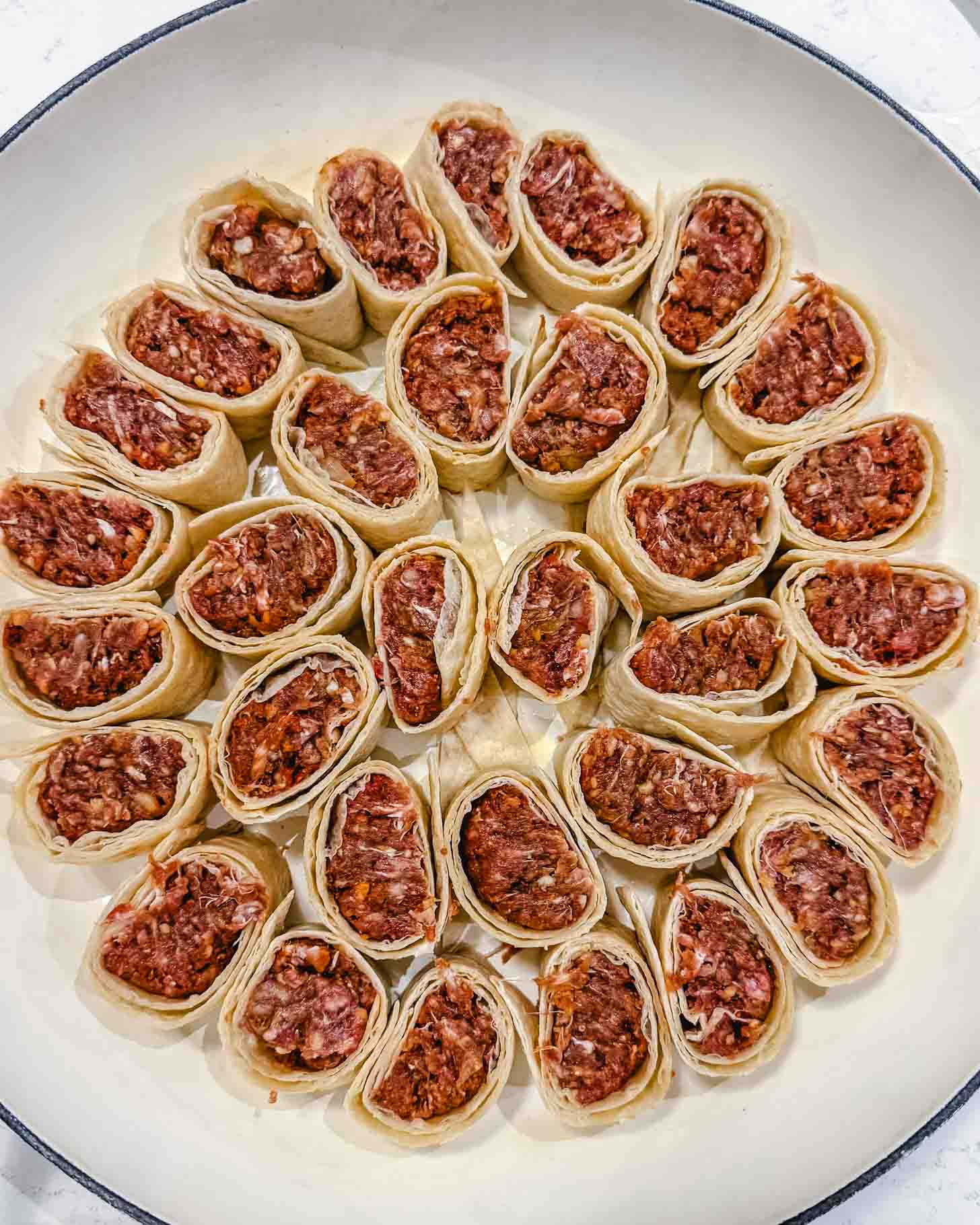 A white round dish filled with neatly arranged pieces of raw pastry rolls, each stuffed with a ground meat filling, placed in a circular pattern, ready to be cooked.