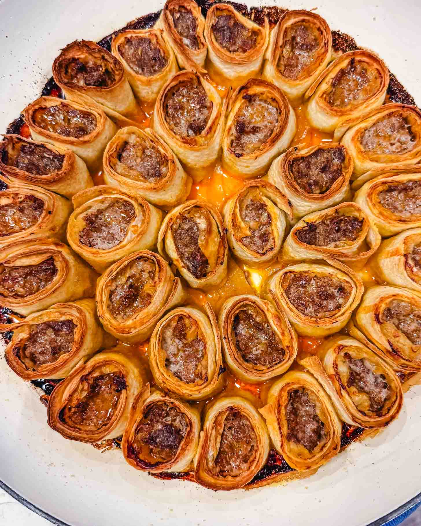A close-up of golden-brown, rolled pastries filled with minced meat, arranged upright in a circular pattern inside a white baking dish with a slightly crispy, caramelized edge.